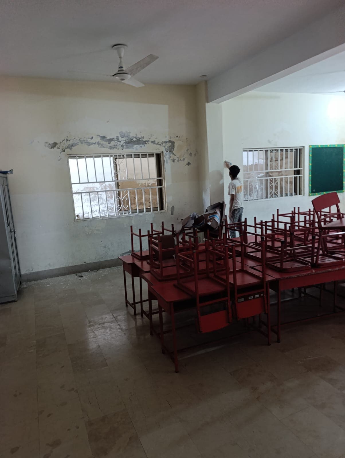 A classroom with peeling, damp-damaged plaster above the window, a painter starting work
