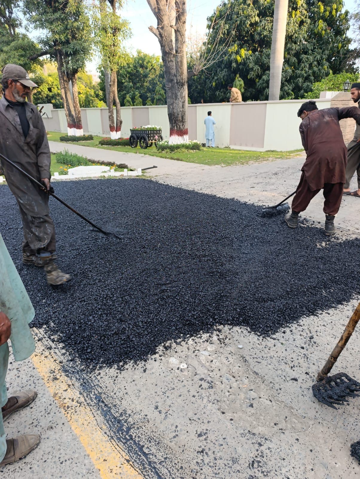 Workers raking hot asphalt into a road patch beside a white boundary wall