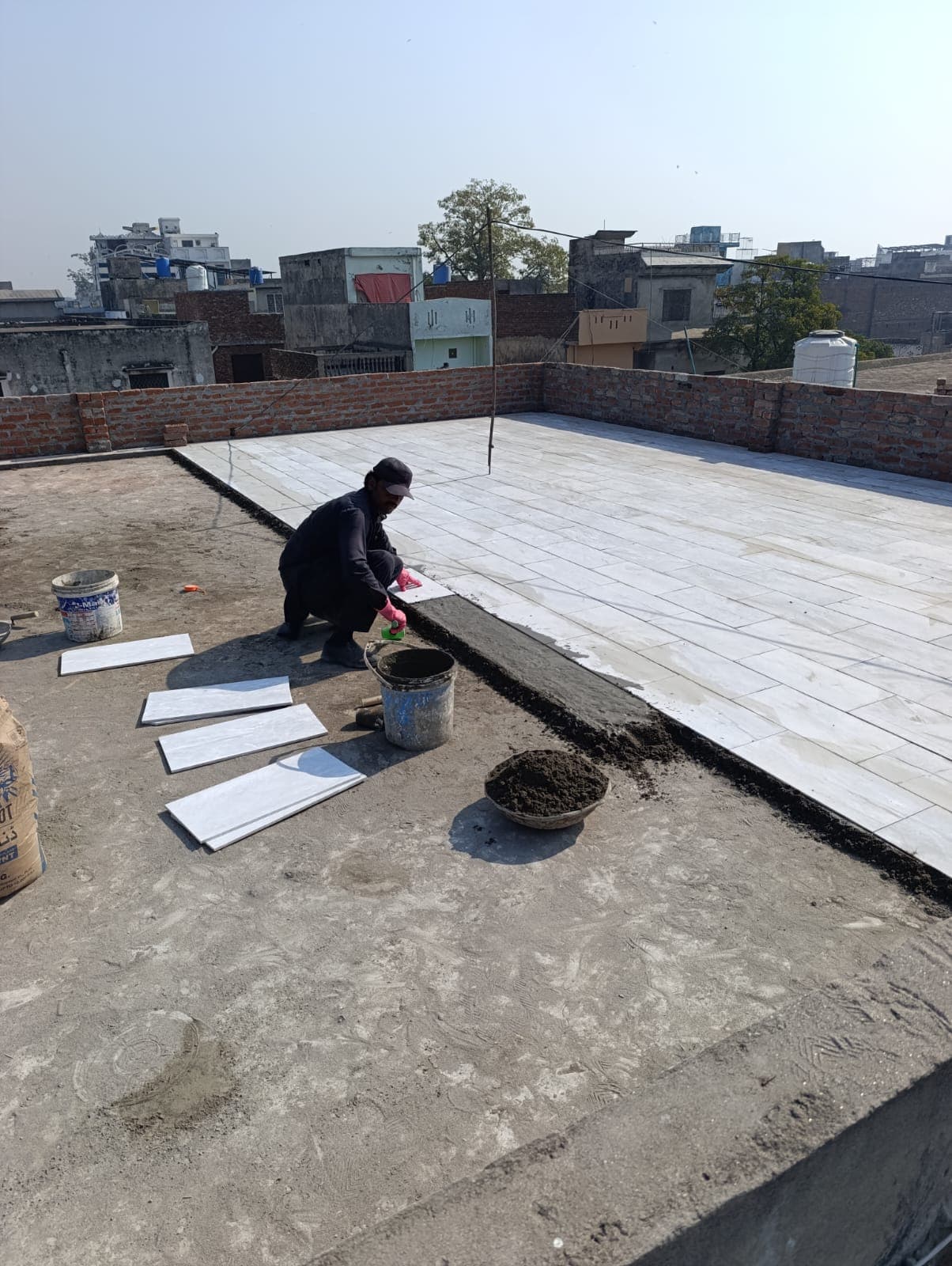 A worker laying white tiles on a mortar bed across an open rooftop