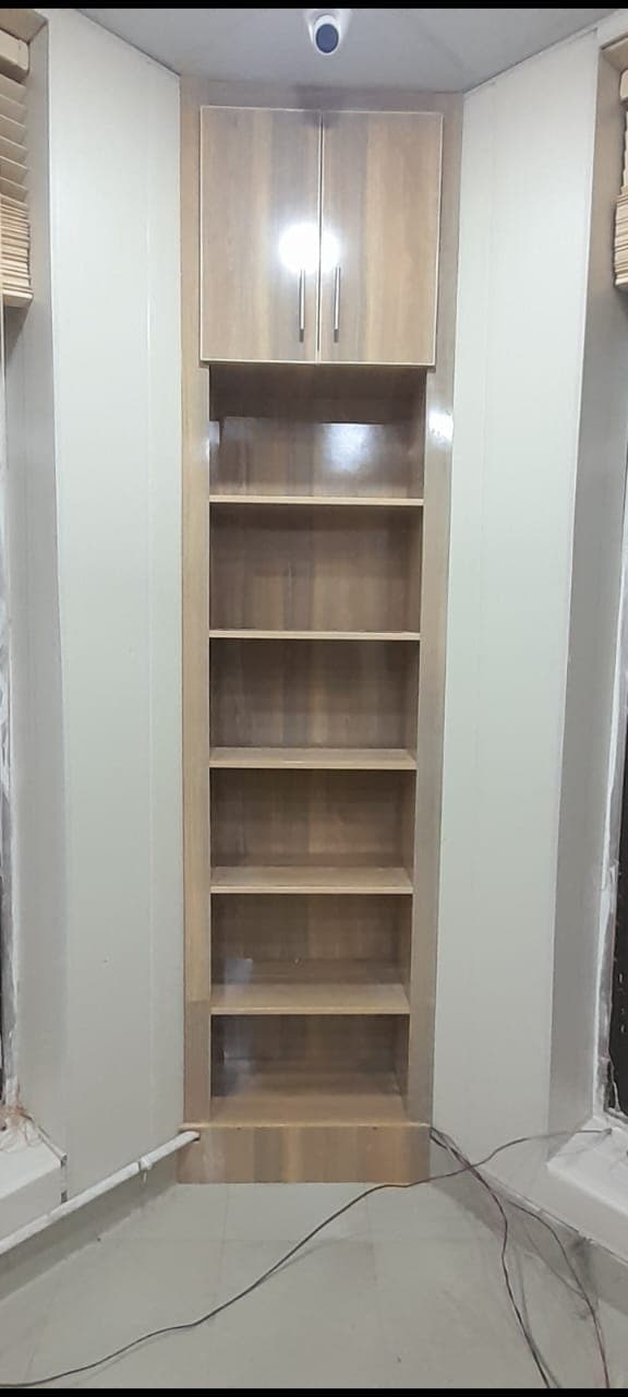 A tall wood-finish cabinet with open shelves and an upper cupboard, fitted into a narrow niche